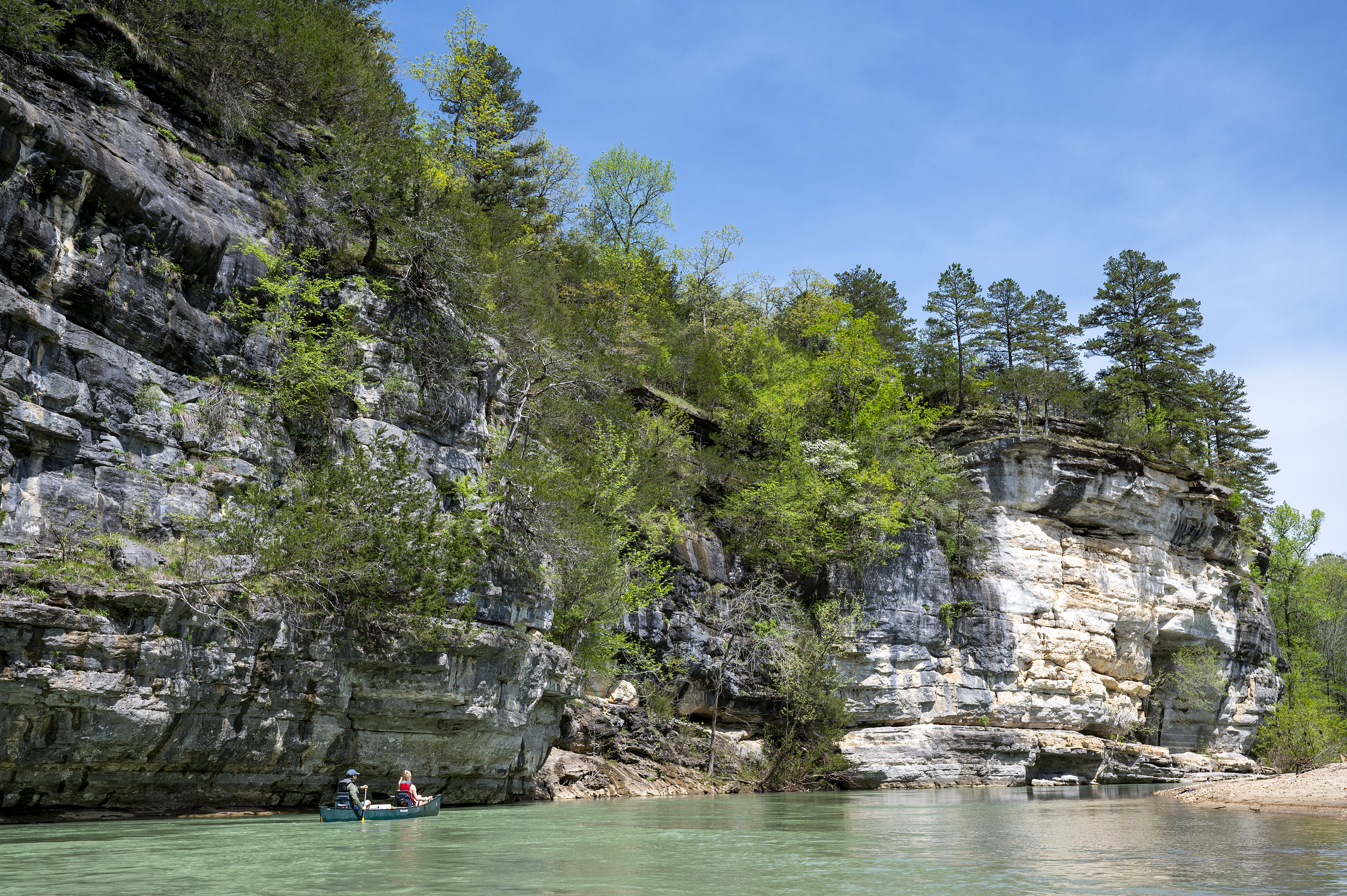 Two people riding a canoe on the Buffalo National River next to a large bluff covered in foilage.