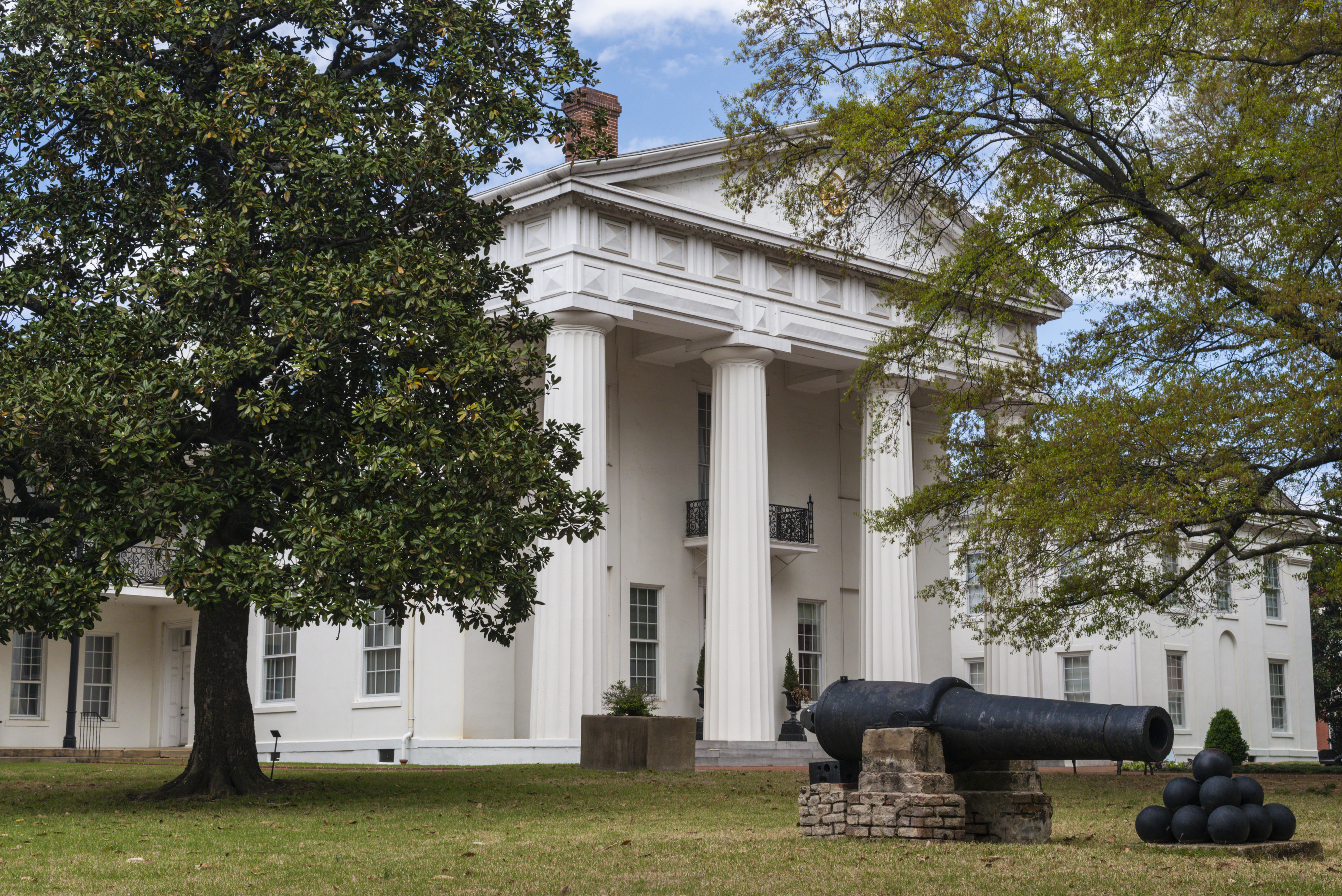 The Arkansas Old State House Museum framed by large trees with a statue of a canon and cannon balls in front.