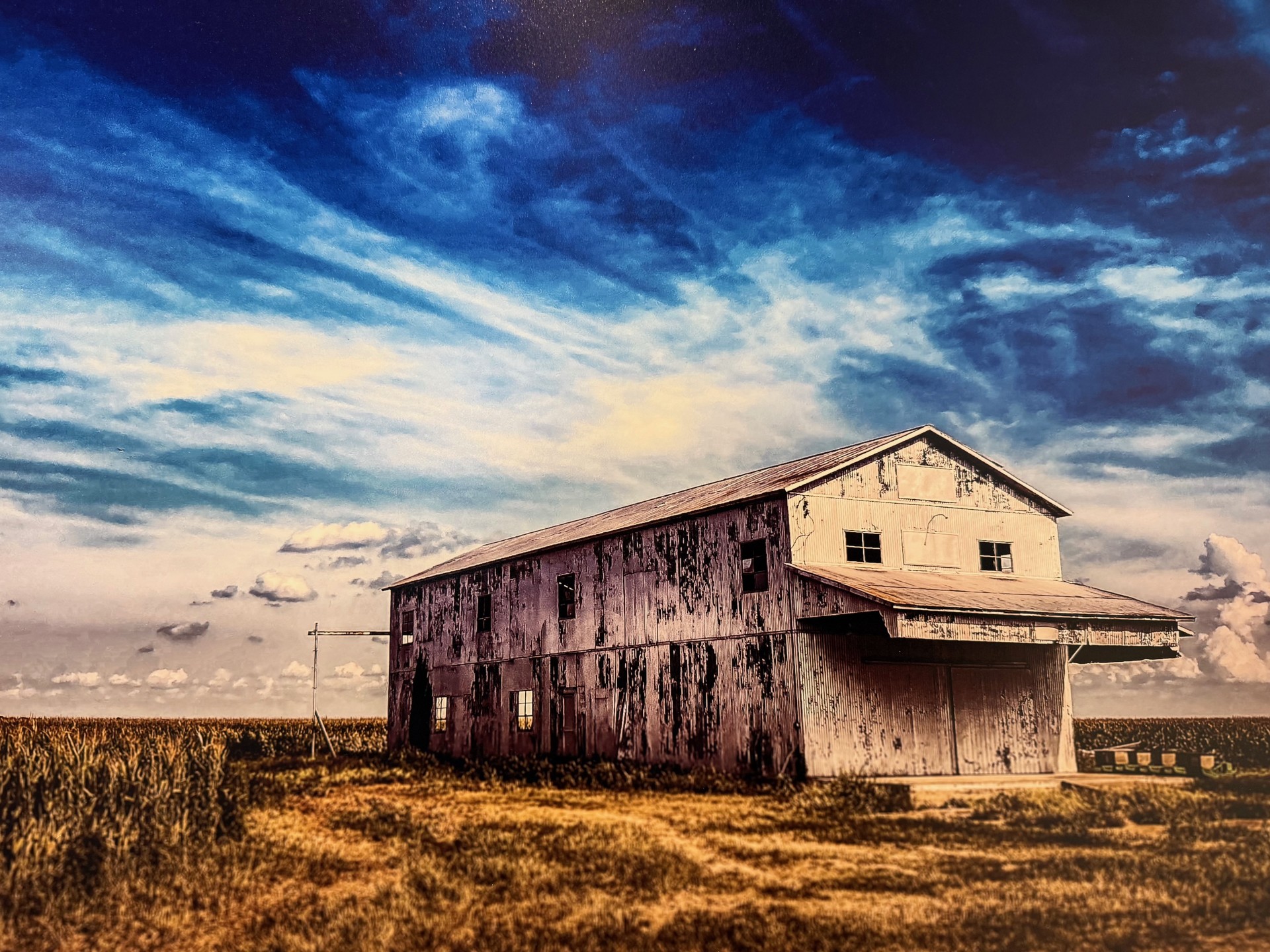 An old white peeling barn in the middle of a field under a bright blue sky covered in wispy clouds.
