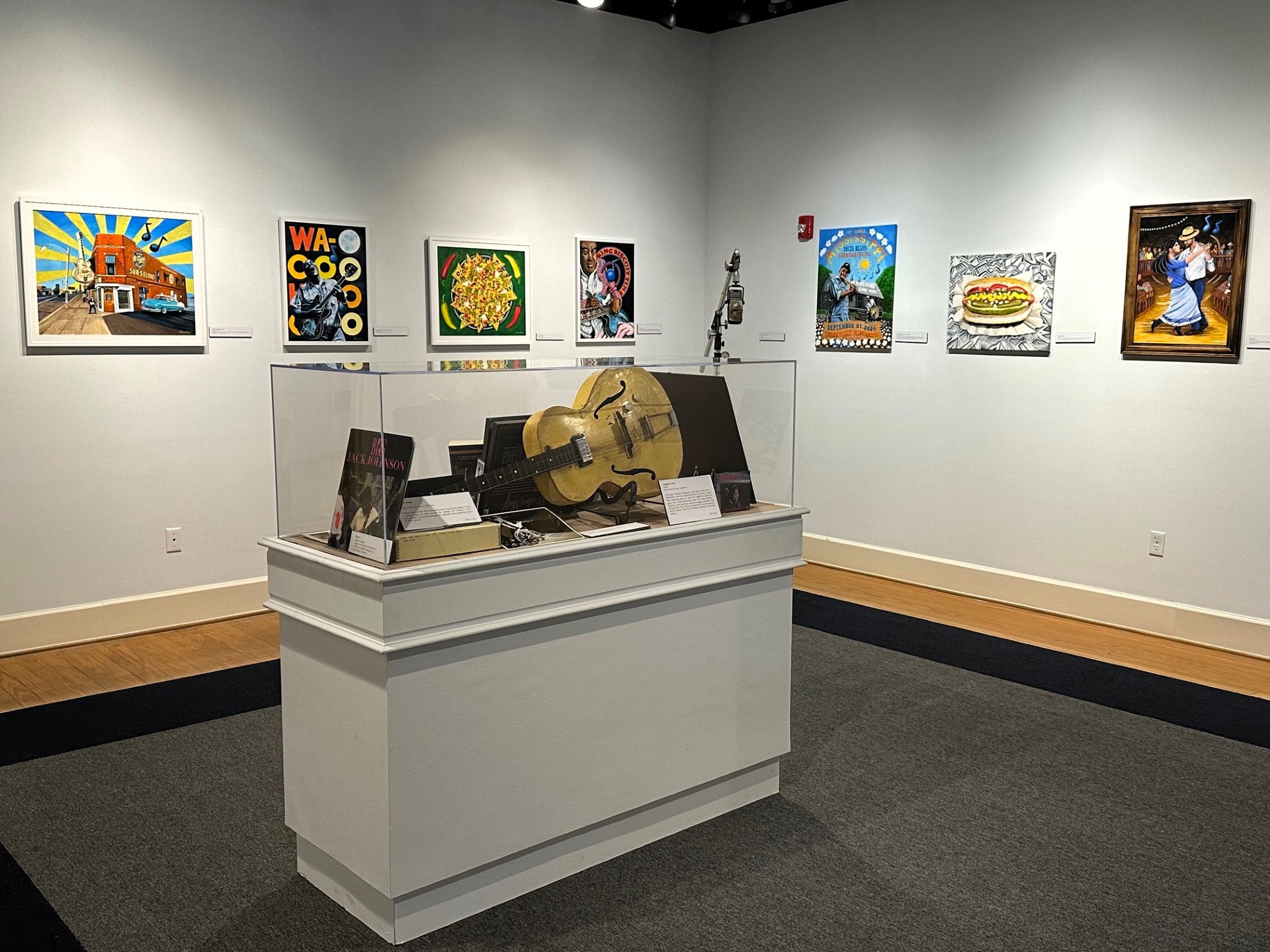 A display case with a guitar laying flat inside is centered in a room with artwork hung gallery style on the walls behind it.