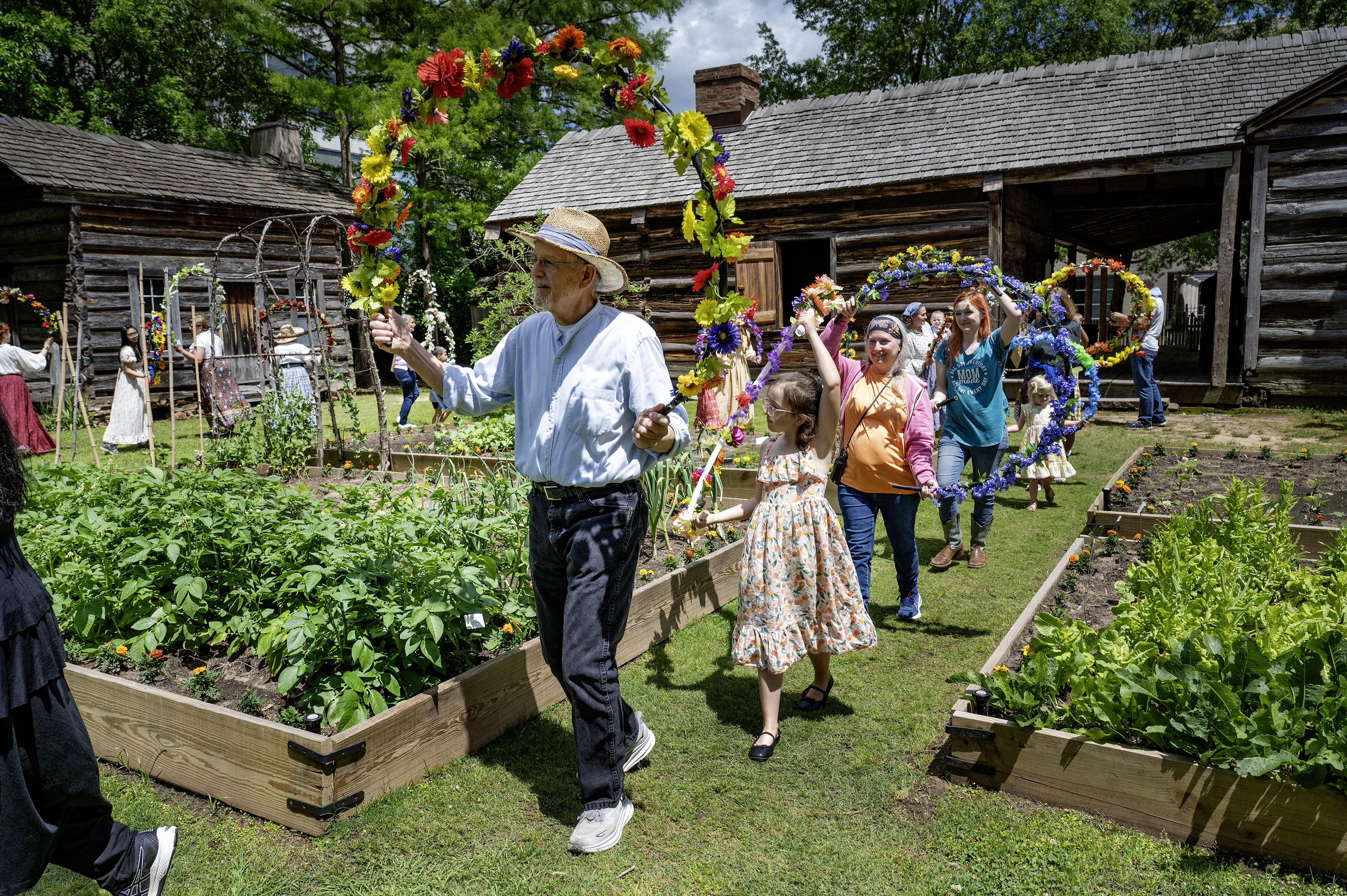 Historic Arkansas Museum Territorial Fair. People walking through garden beds in a line holding hula hoops decorated with flowers.