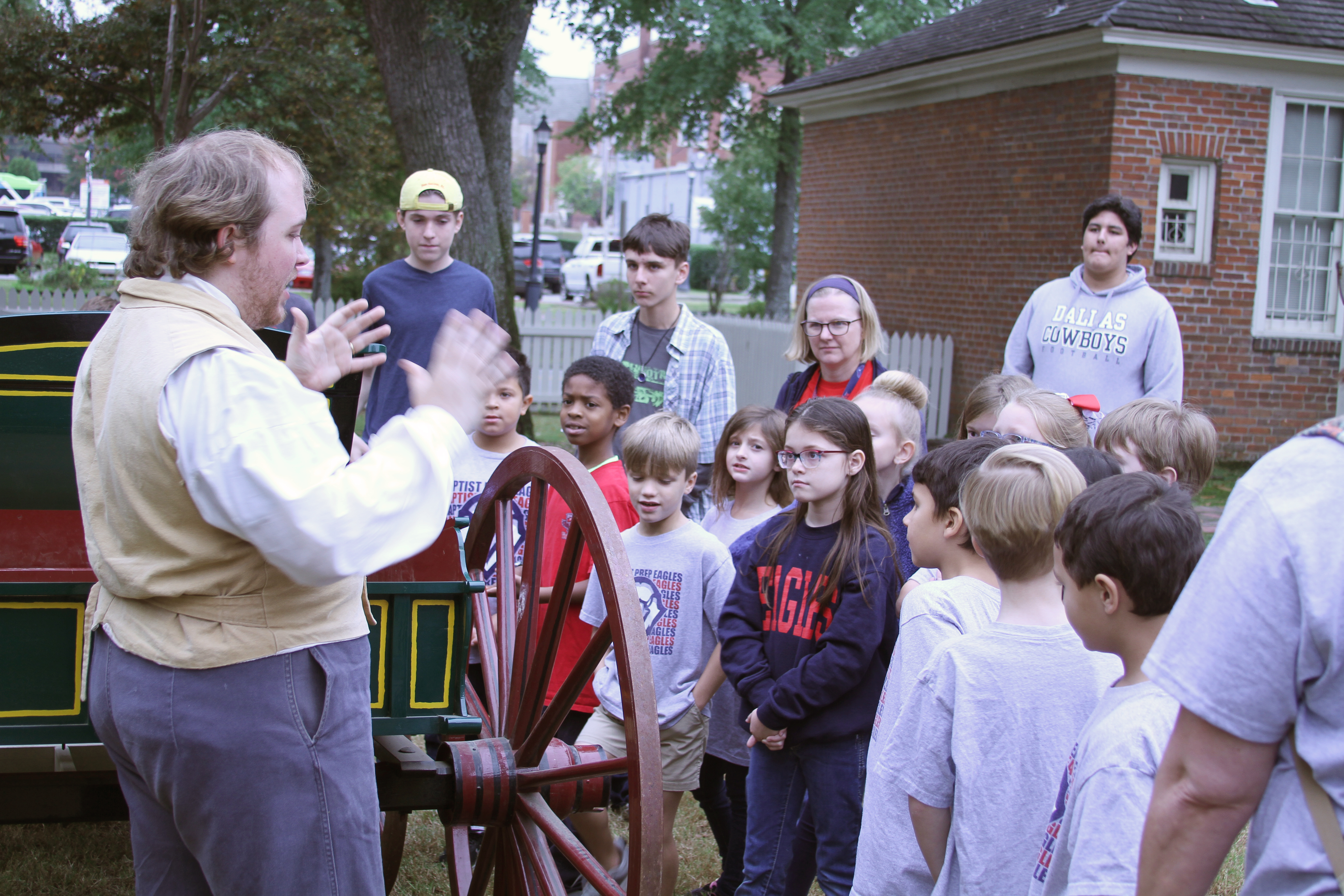 A man in period dress standing next to a older style wagon speaking to a group of children.