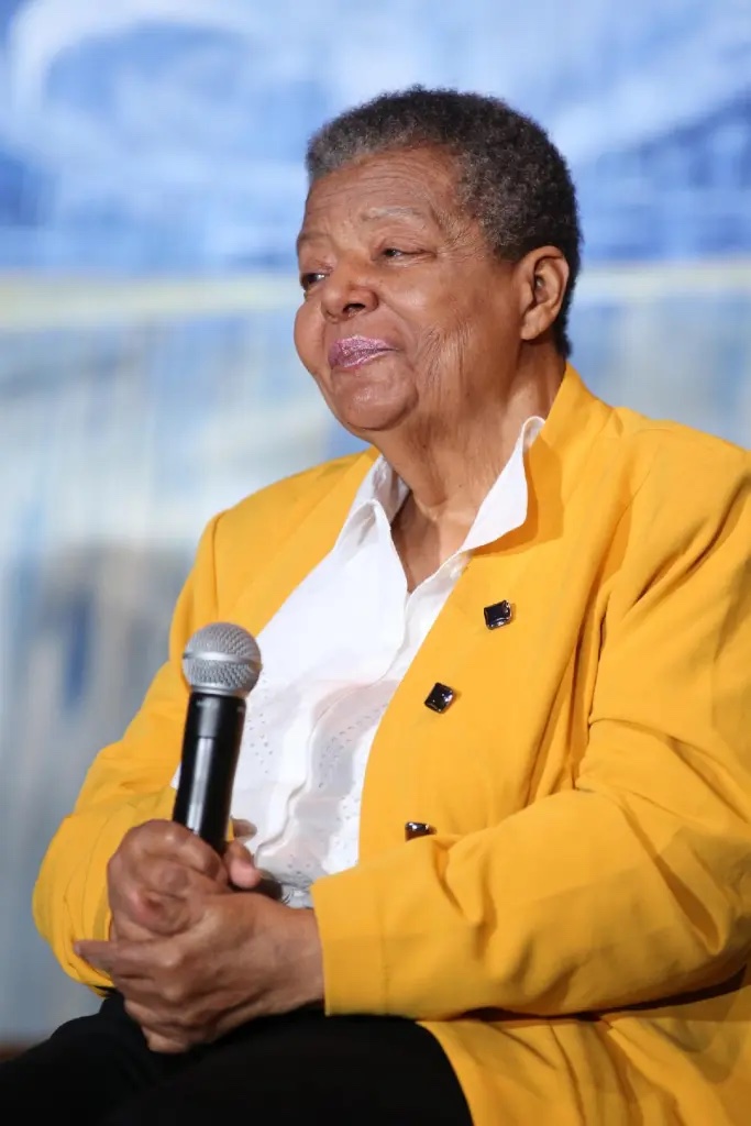 An older woman in a yellow blazer and white blouse seated with a microphone on a stage.