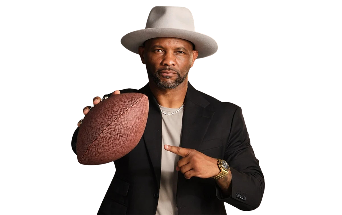 A man in a black blazer and a gray hat holds a football in his right hand and points to it with his left.