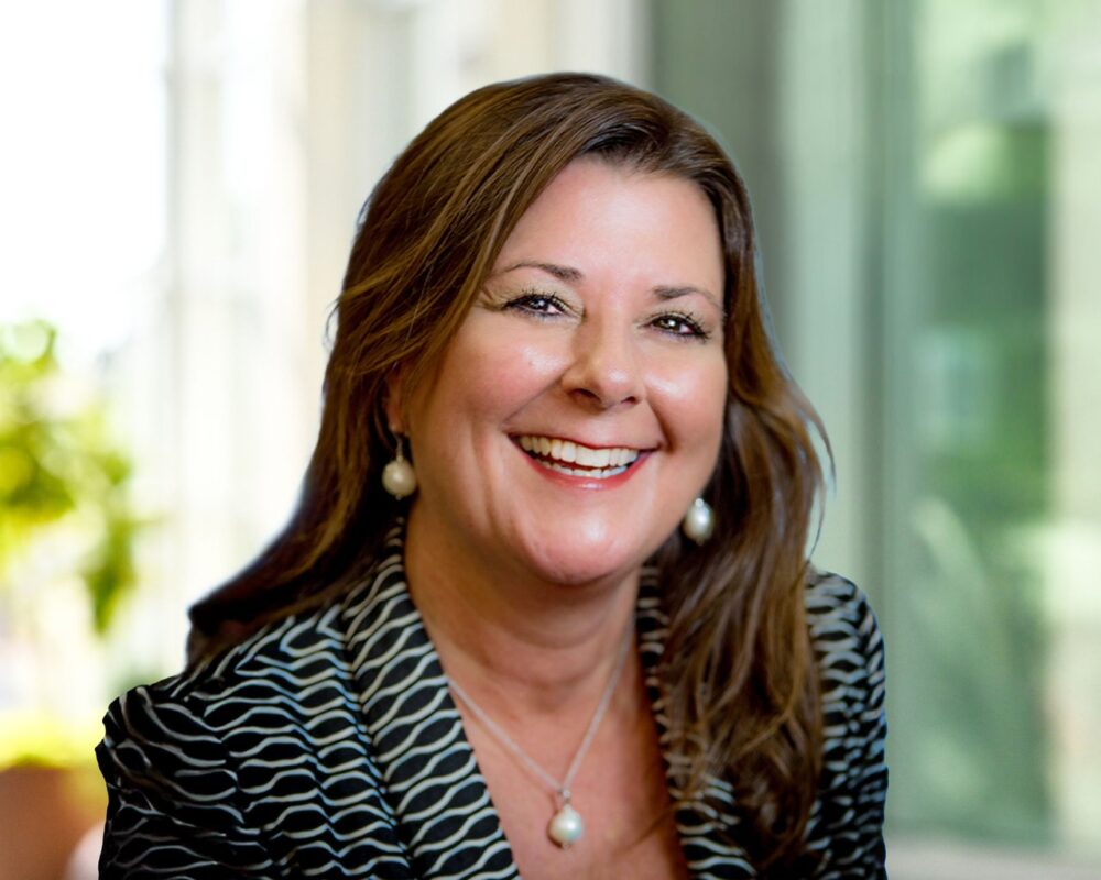 Gina Gimberling, a woman in a black and white blazer and large pearl earrings with medium long brown hair smiling.