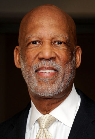 An older man dressed in a black suit with a yellow tie smiling.