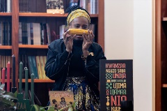 A Black woman holds up an ear of corn. She is standing next to a Kinara and behind a poster for Kwanzaa