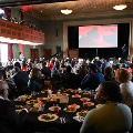 Alargegroupofpeoplesittingatcirculartableswatchingapresentationontheballroomstage